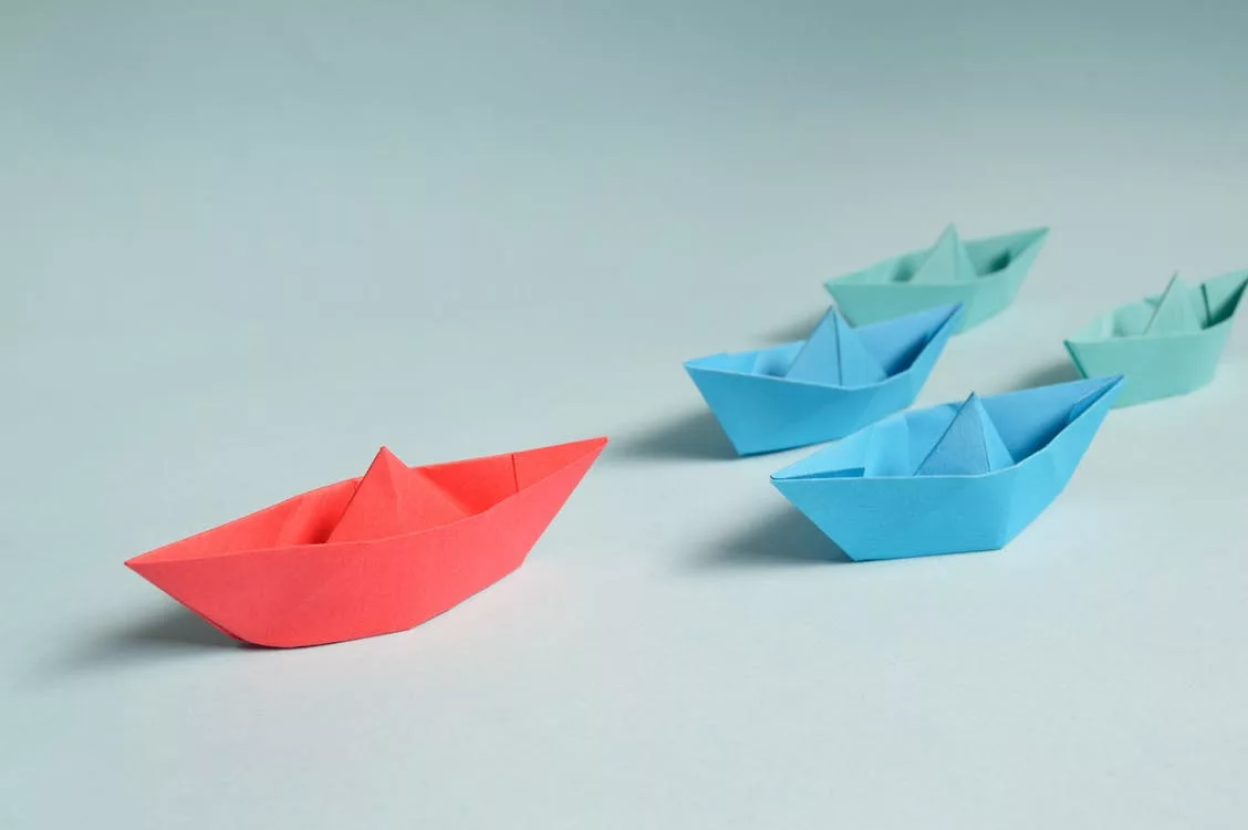 Red paper boat leading blue paper boats on a light blue background