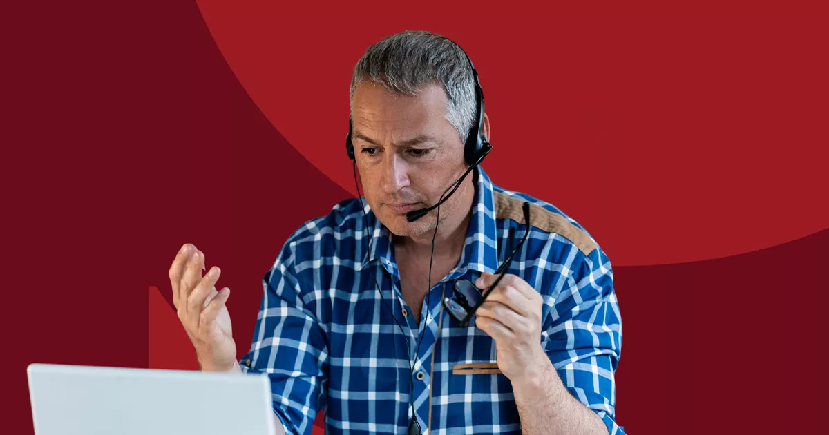 Man in blue plaid shirt with headset having video call on laptop against red background.