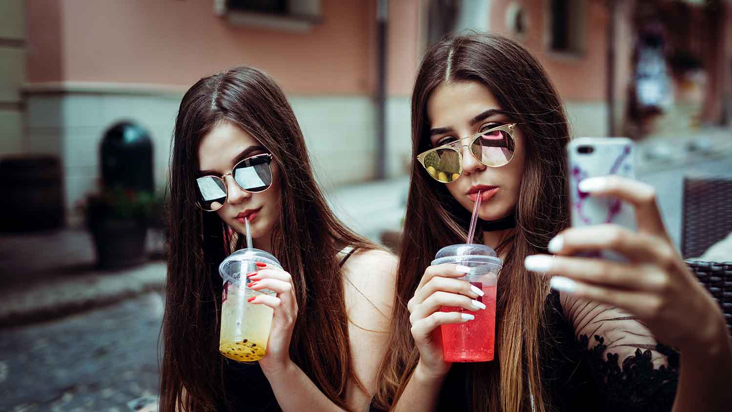 Two women in sunglasses sipping iced drinks and taking a selfie on a sunny street.