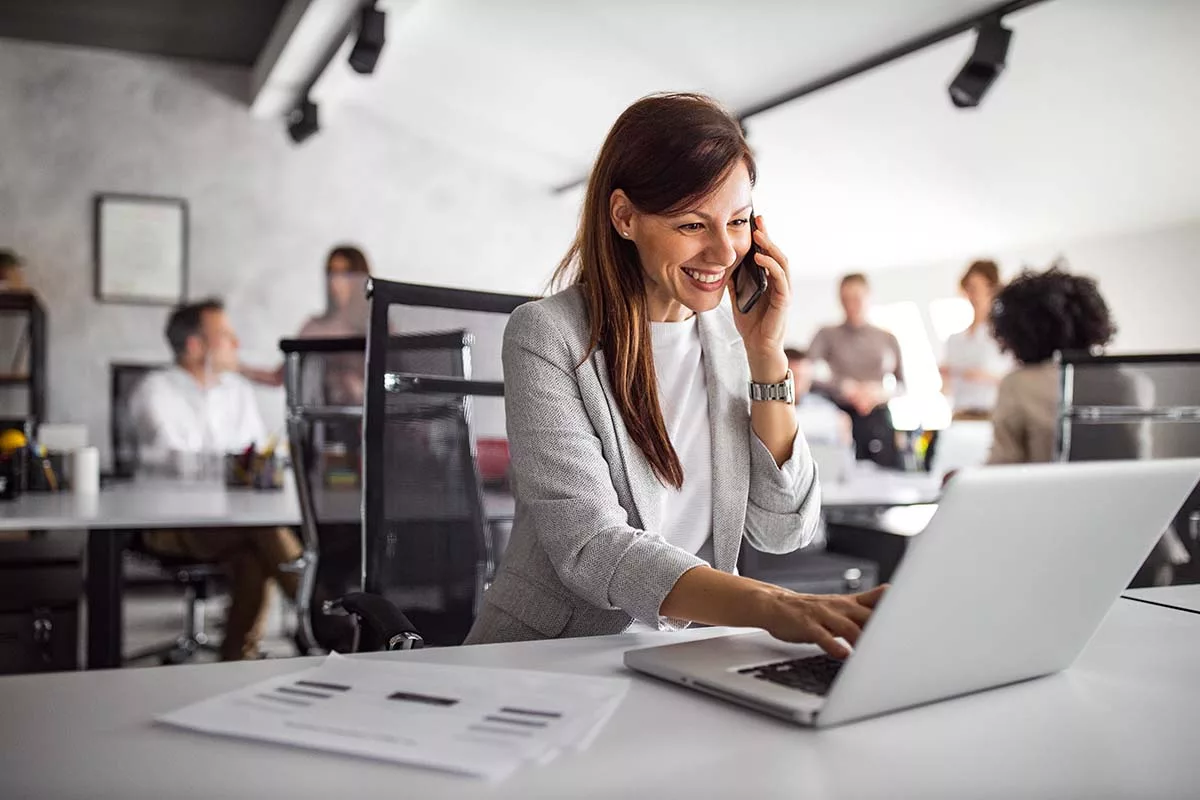 Smiling businesswoman on phone using laptop in modern office