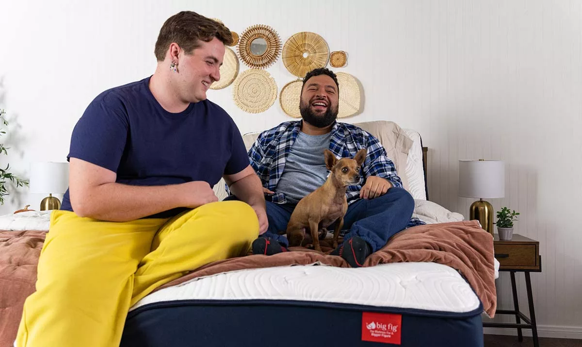 Two men smiling on a bed with a small dog