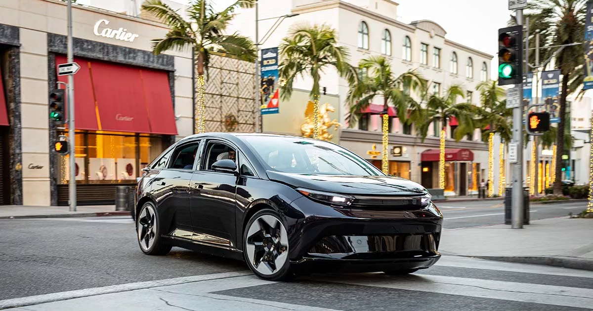 Sleek black electric car driving through a city street lined with palm trees and luxury stores.