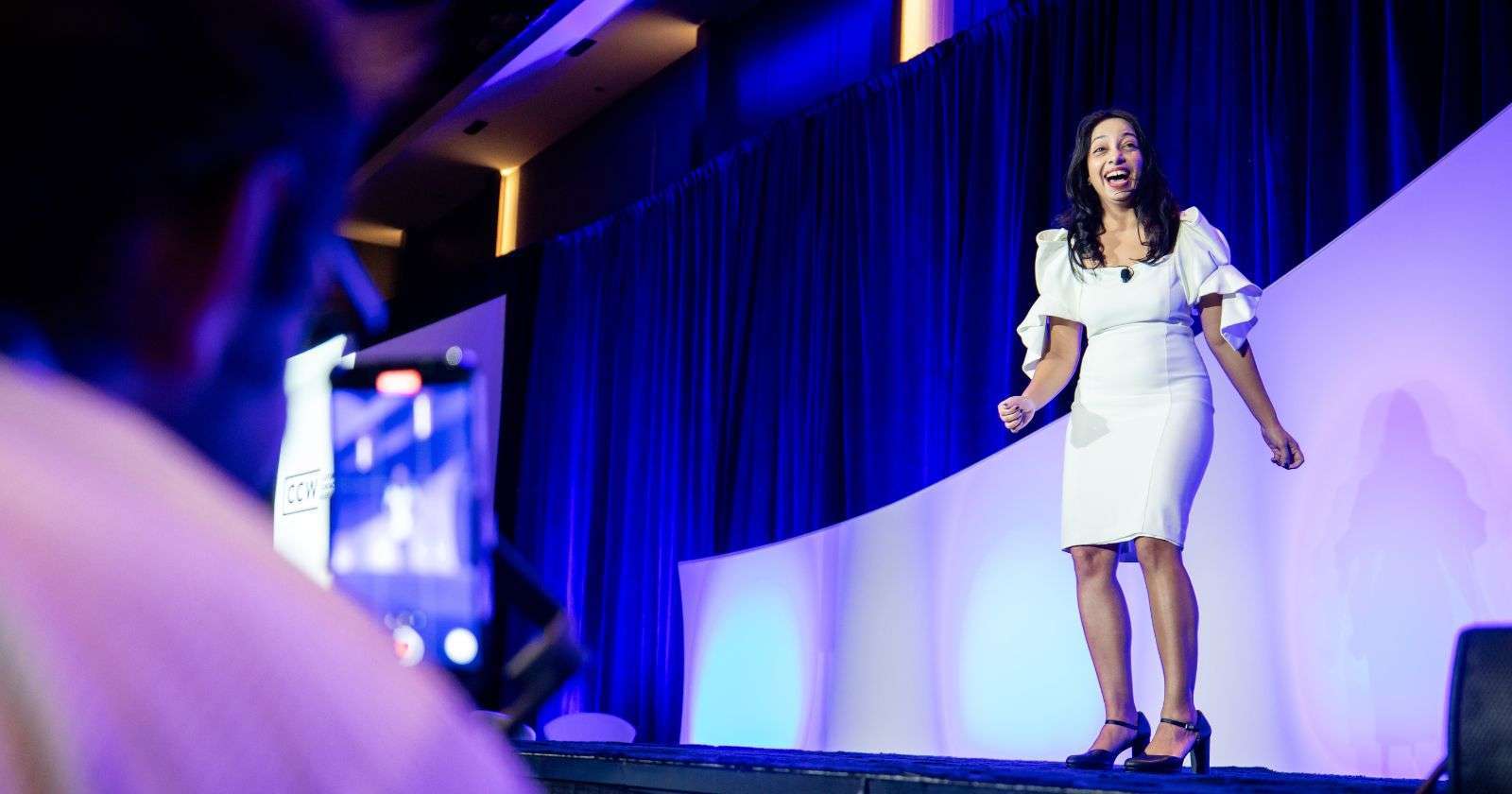 Woman in white dress speaking energetically on stage