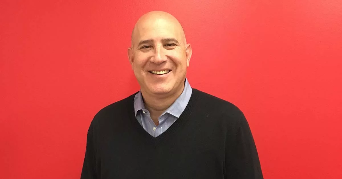 Smiling man in a black sweater and blue shirt against a vibrant red background.