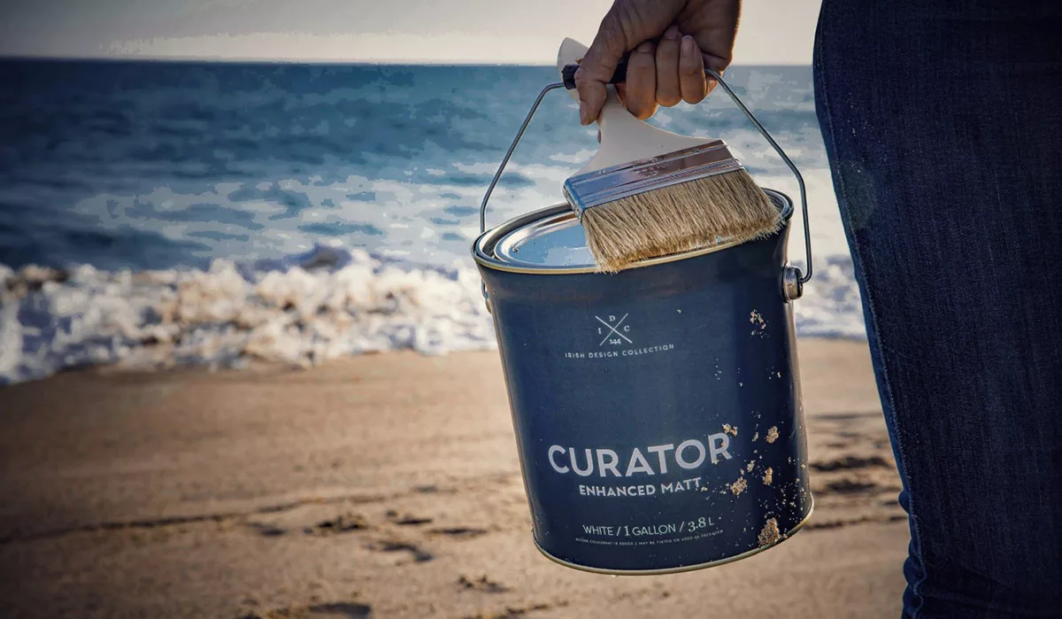 Hand holding paint can and brush on sandy beach with ocean waves in the background.