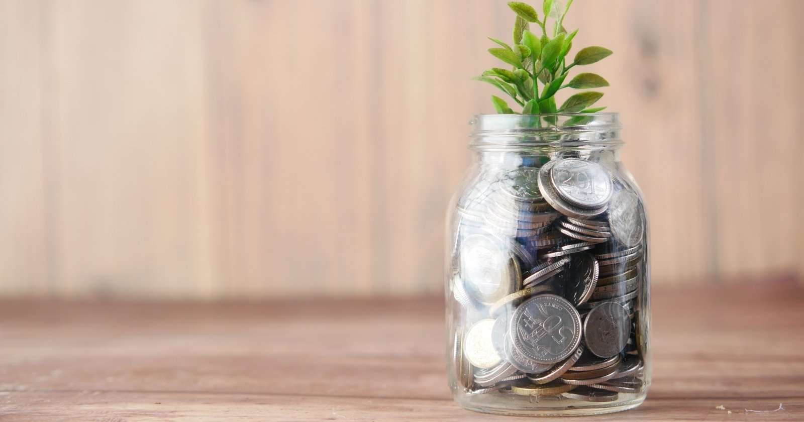 A mason jar of coins with a small plant growing out of the top against a wooden background.