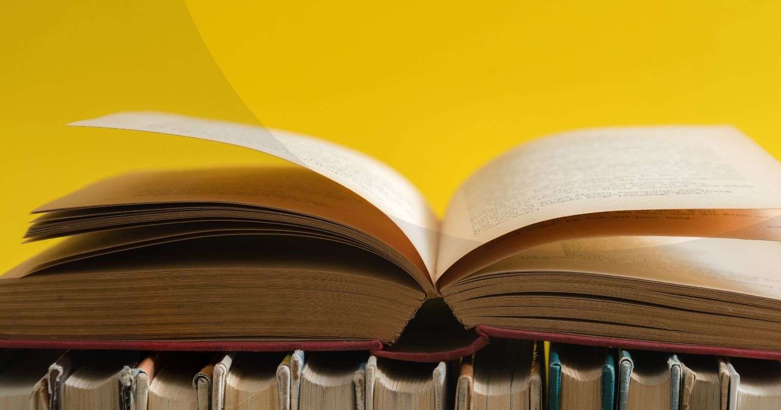 An open hardcover book resting on top of other hardcover books against a yellow background.