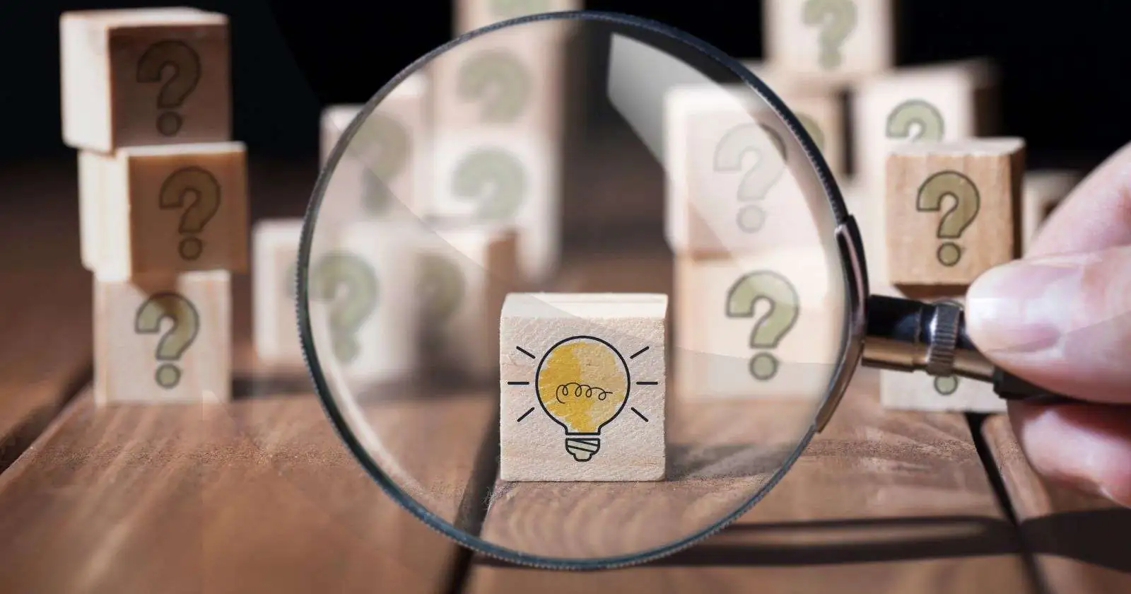 Magnifying glass hovering over a wooden block with a light bulb on it with other wooden blocks with questions marks on them behind it.