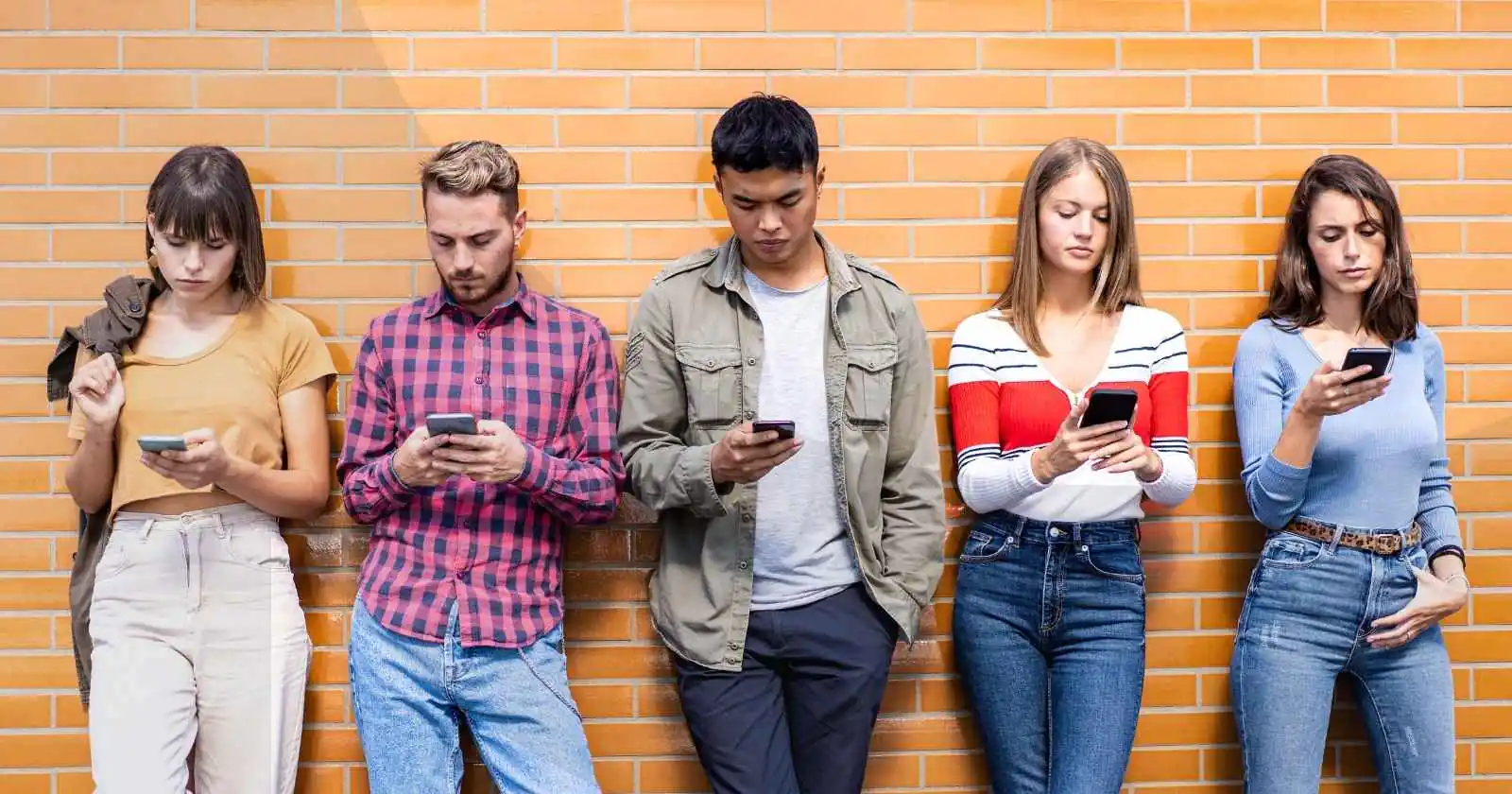 Five young adults using their cell phones while leaning against a brick wall.