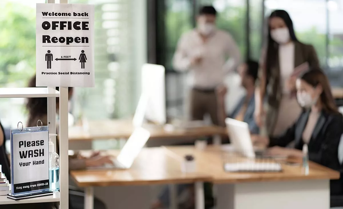 Office reopening with masked employees and social distancing sign. Hand wash reminder at workspace entrance.