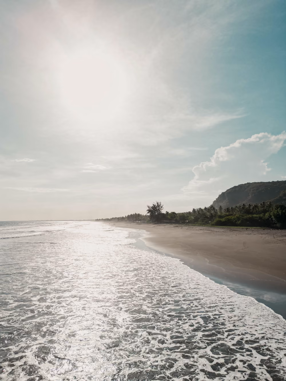 Sun shining over a beach with foamy waves, green trees, and a mountain in the distance.