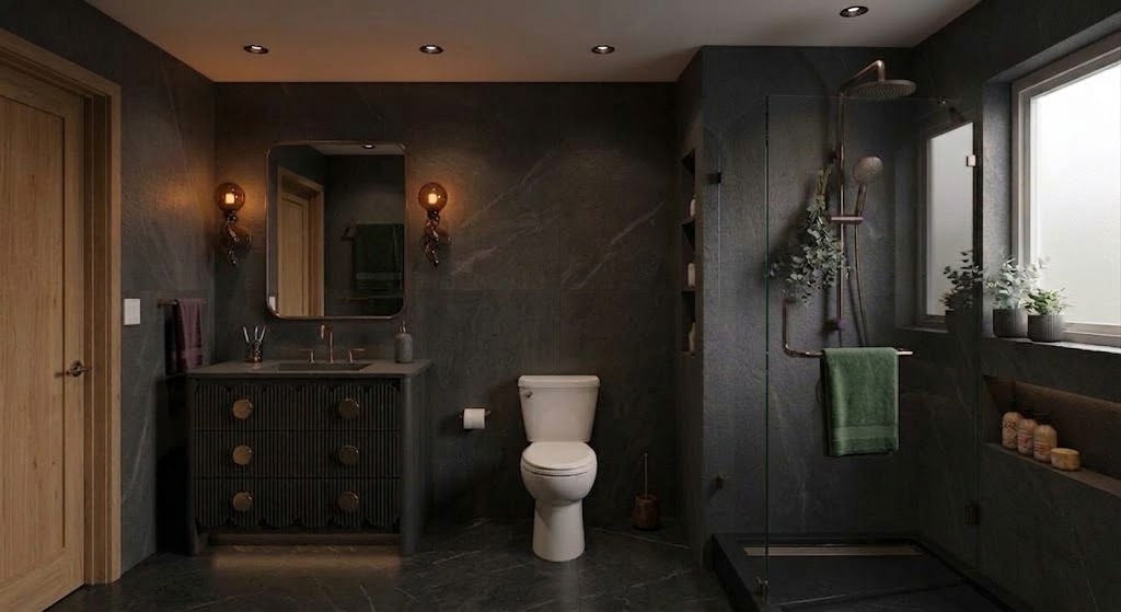 Modern bathroom with dark stone walls and floor, wooden door to the left, a vanity with round brass knobs and a mirror above it lit by two amber wall sconces, a white toilet in the center, and a glass-enclosed shower with a rain showerhead, green towel, built-in shelves, and window with potted plants on the right.