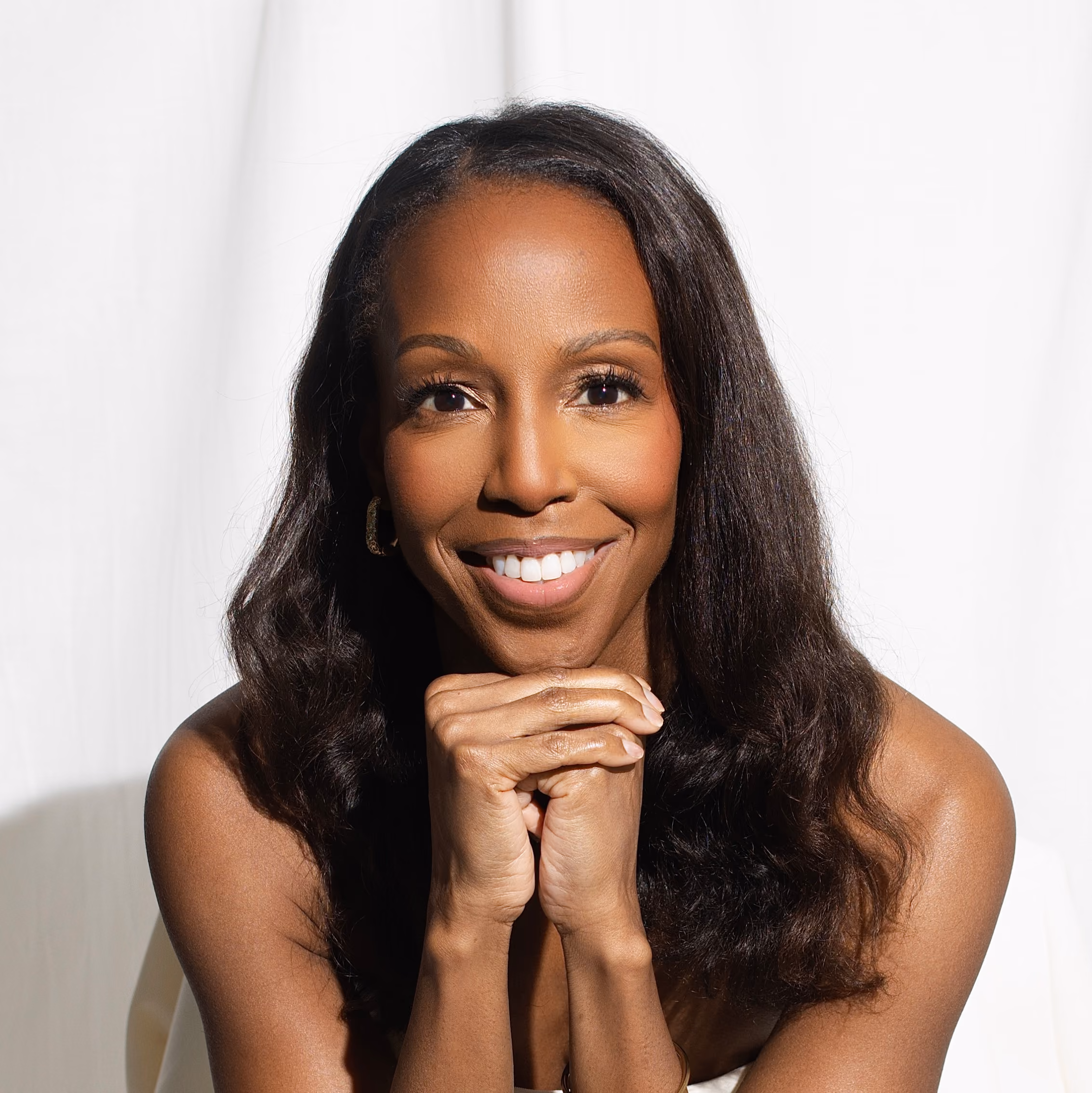 Portrait of Sarah Lewis, smiling with her chin resting on her clasped hands, against a soft white backdrop.