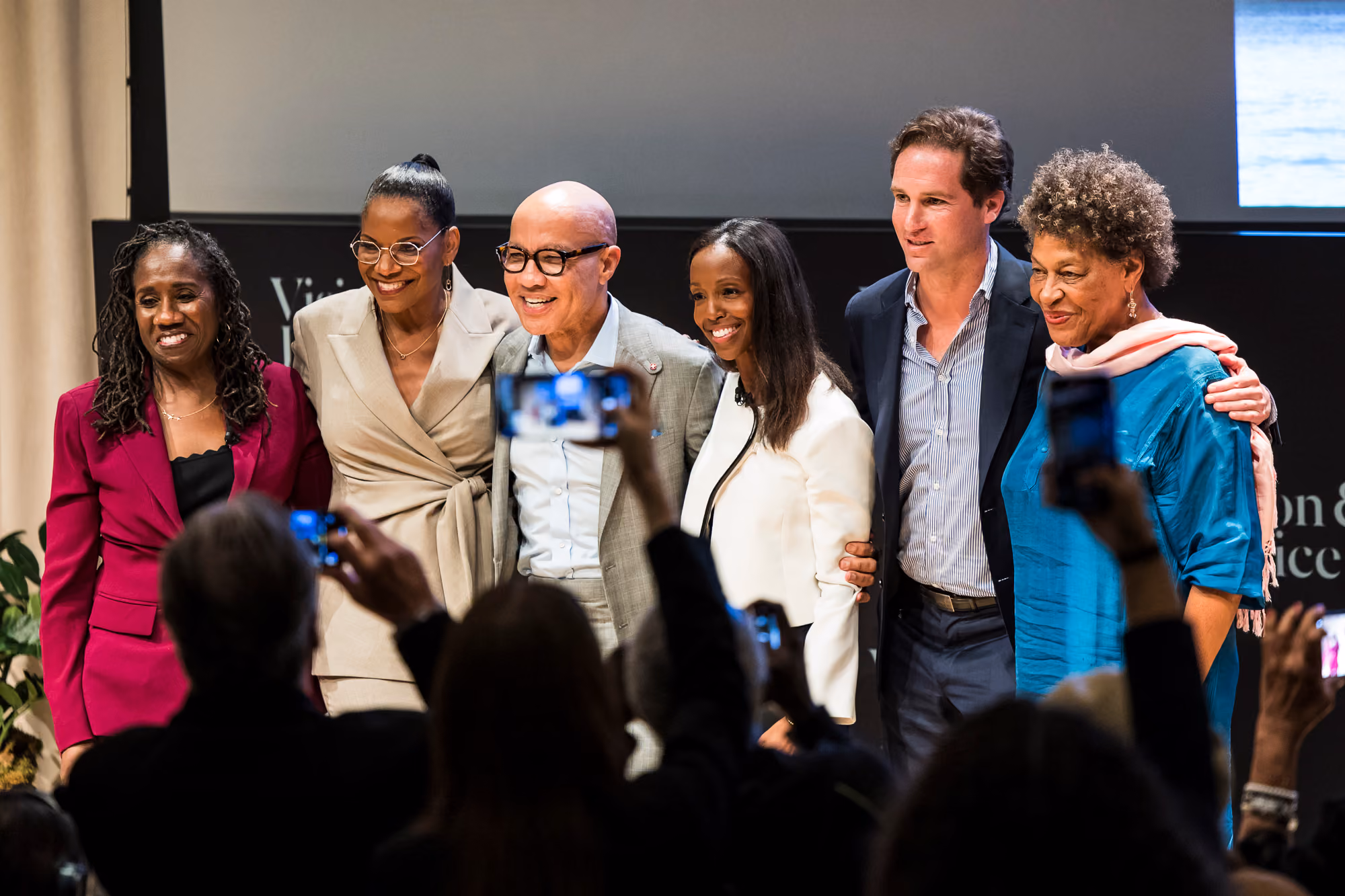 A group of six people stands before a crowd while being photographed on stage.