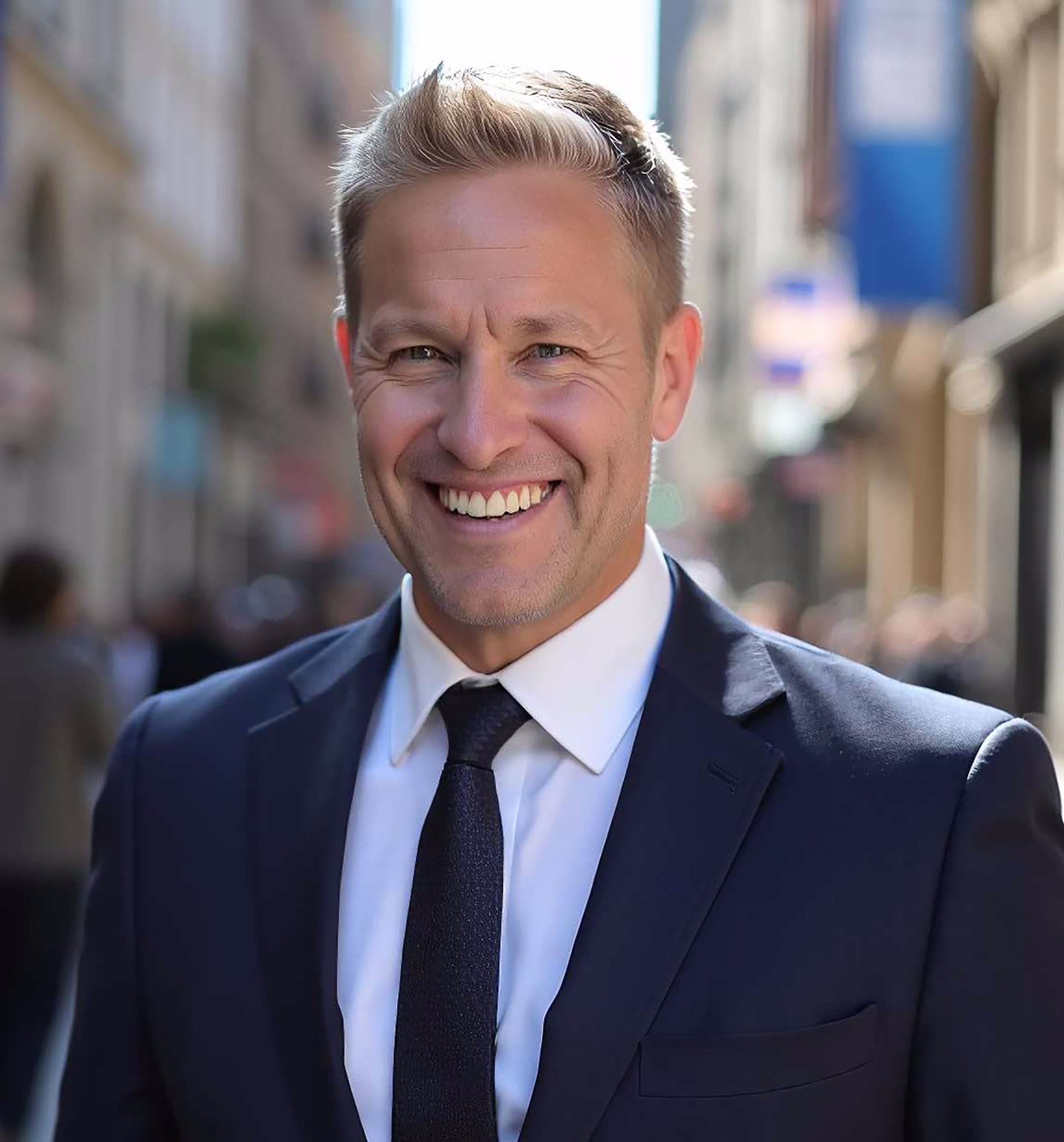 Smiling man in a dark suit and tie standing on a city street with blurred buildings behind.