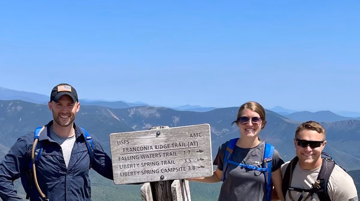 Three hikers smiling beside a wooden trail sign with mountain range in the background.