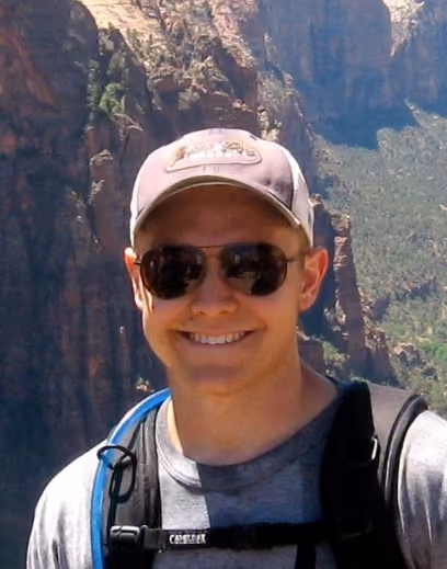 Smiling man wearing sunglasses, a cap, and a backpack standing in front of rocky mountains.