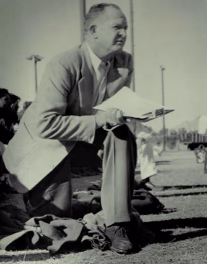 Man in a suit kneeling outdoors holding papers, with a sports field and players in the background.
