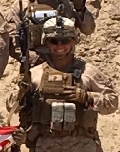 Smiling soldier wearing tactical gear and helmet in a desert setting holding a small box.