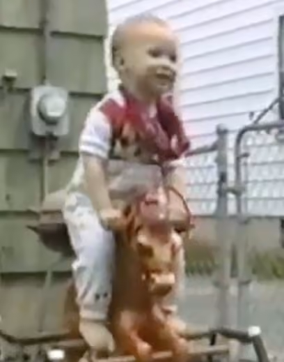 A toddler sitting and smiling on a toy horse outside near a fence and house wall.