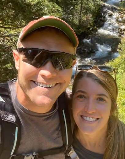 Smiling man and woman outdoors with greenery and a flowing river in the background.