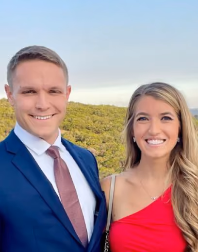 Smiling man in a suit and woman in a red dress posing outdoors with greenery and sky behind them.
