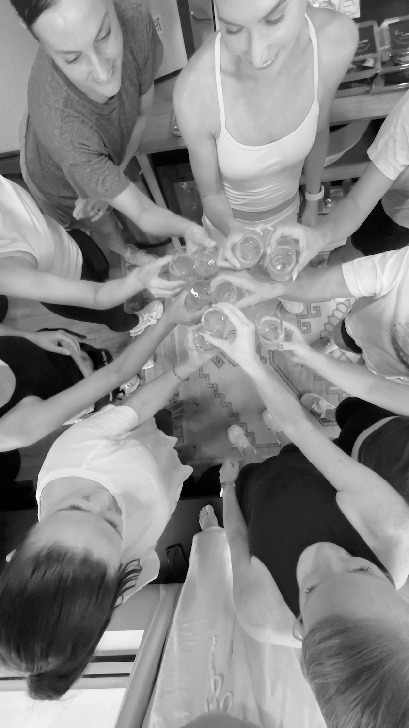 A high-angle, black-and-white photo of a group of people standing in a circle, bringing their clear glasses together in a toast.