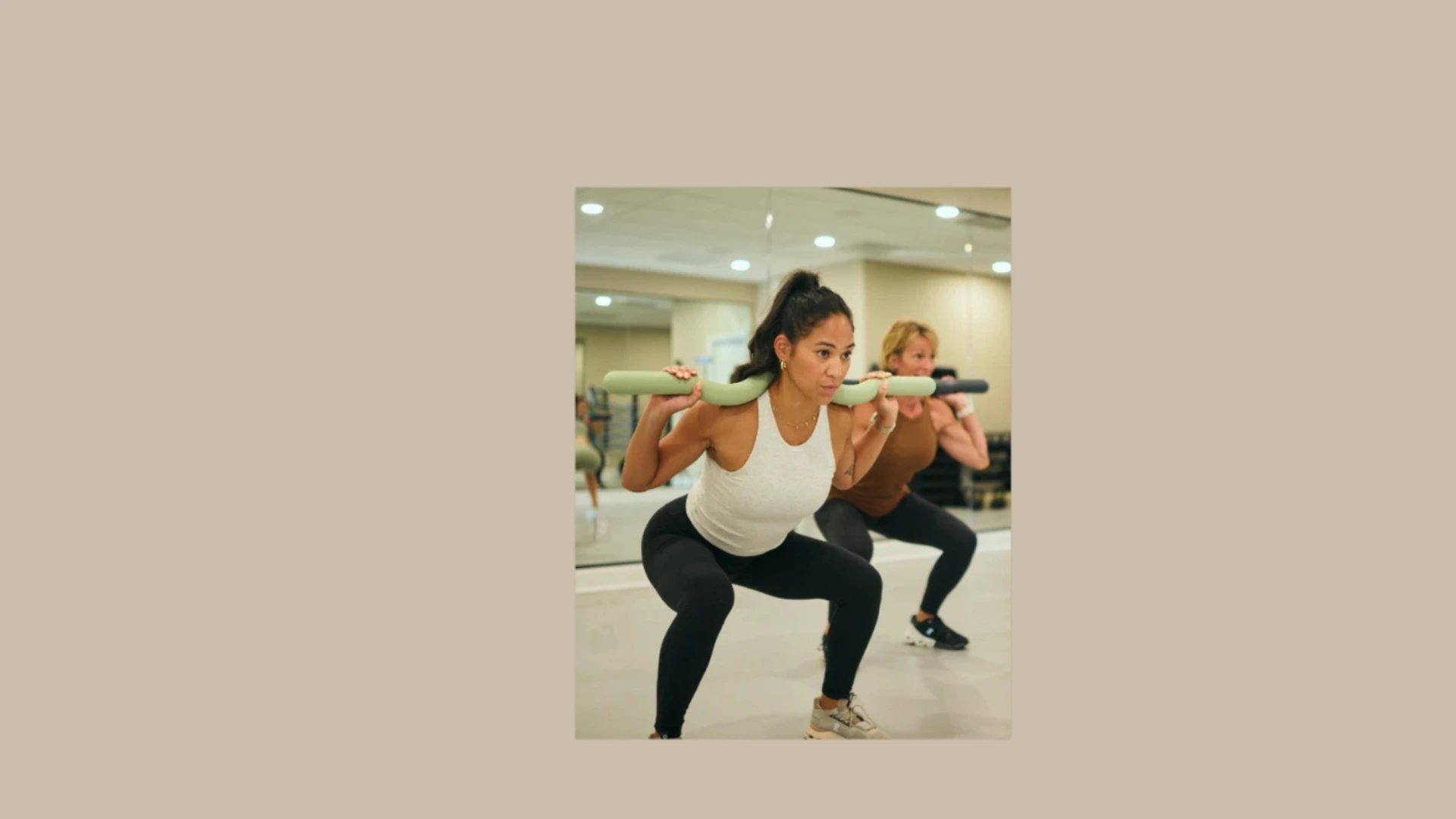 Two women performing weighted squats in a fitness studio class, with a padded exercise bar resting across their shoulders.
