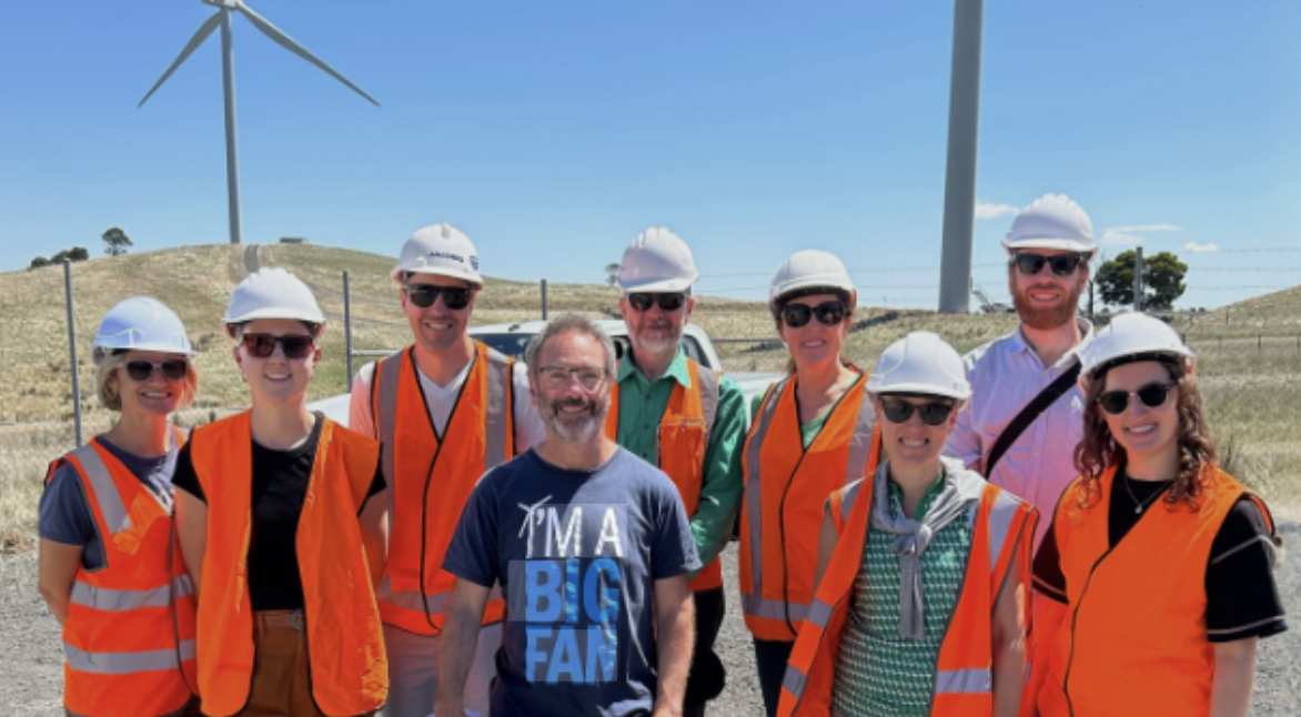 A group of people wearing high-visibility vests and hard hats on a sunny day at a wind farm site, with wind turbines visible in the background.
