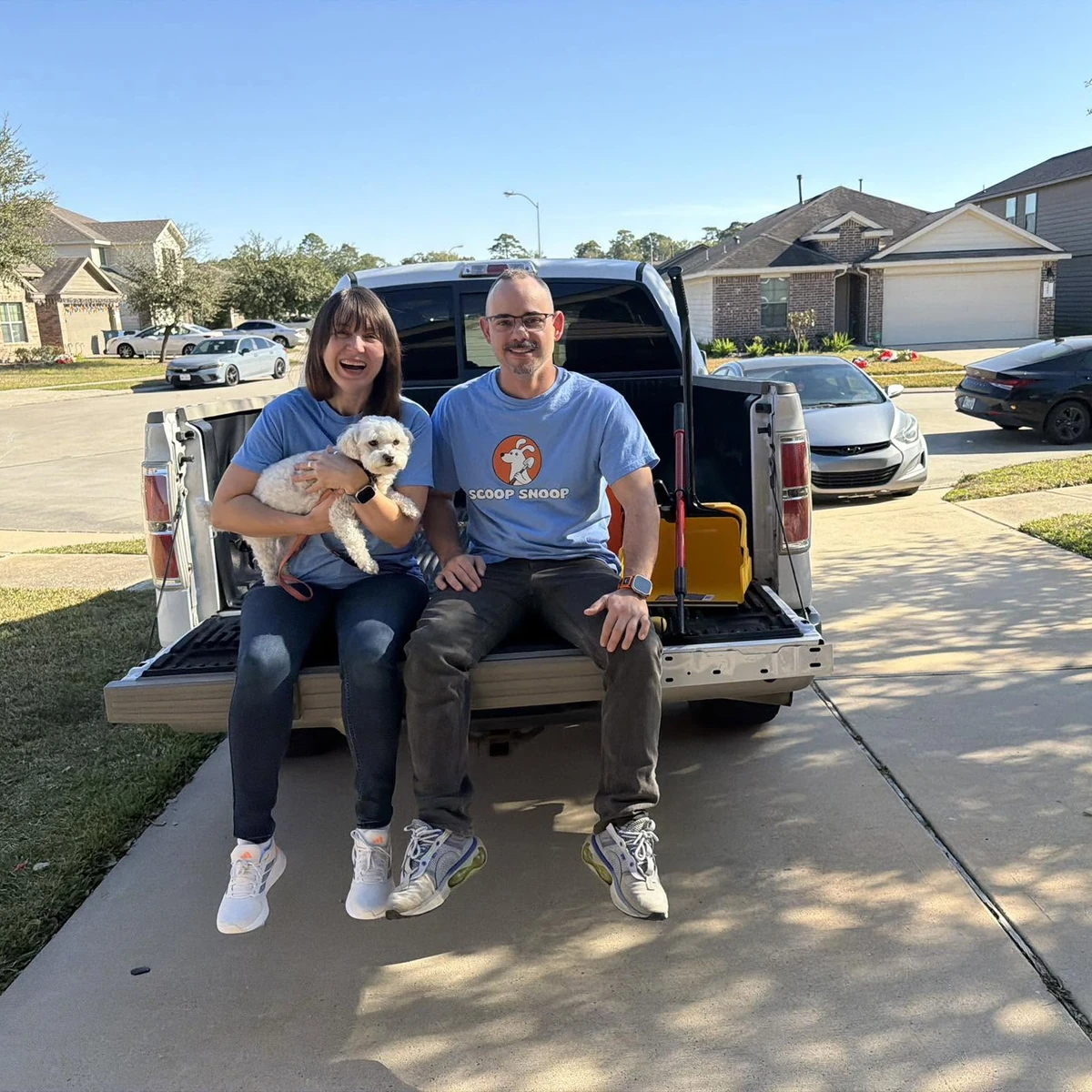 Scoop Snoop pet waste removal team sitting on the tailgate of a work truck with a small white dog in a residential neighborhood.