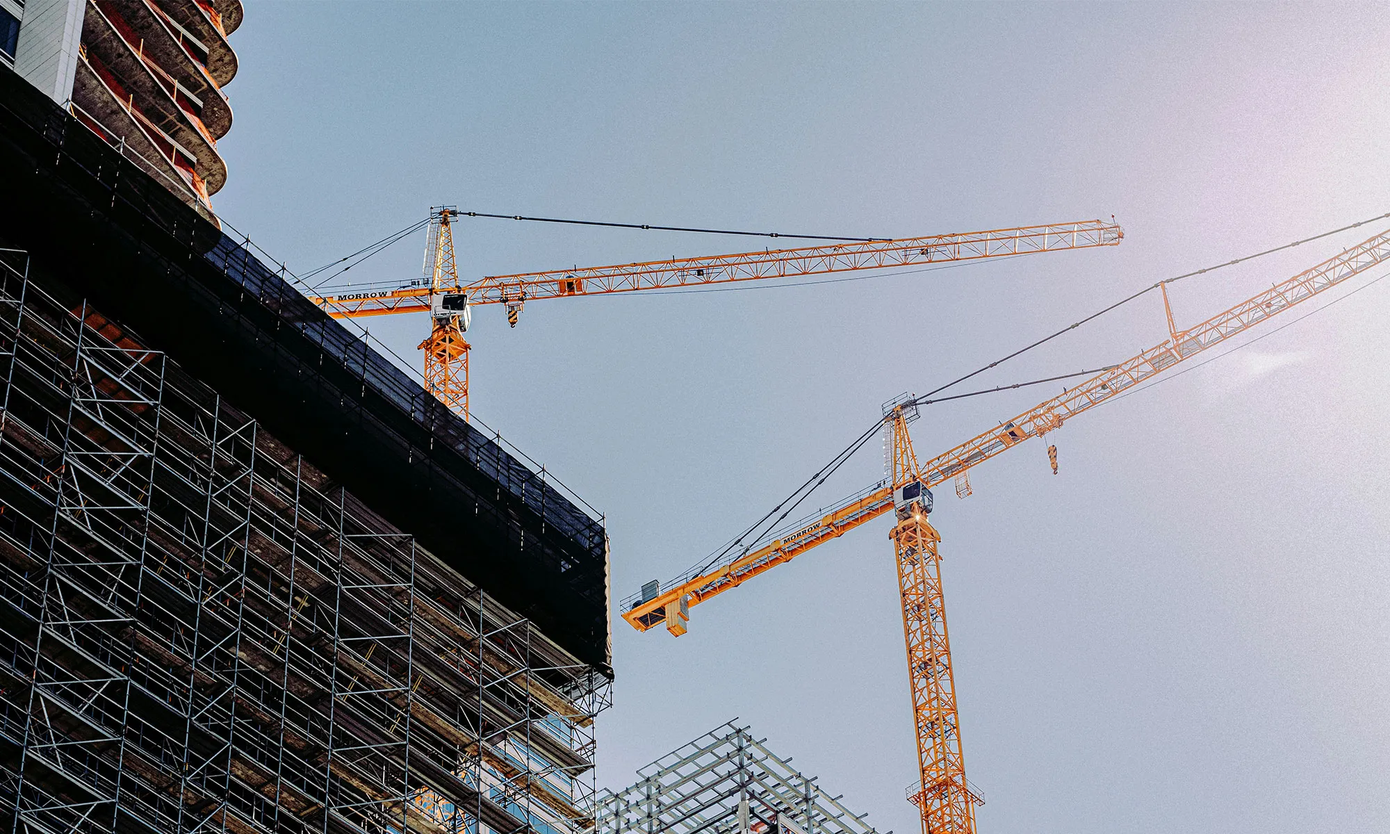 Two tall orange construction cranes over scaffolding on a building under construction against clear sky.