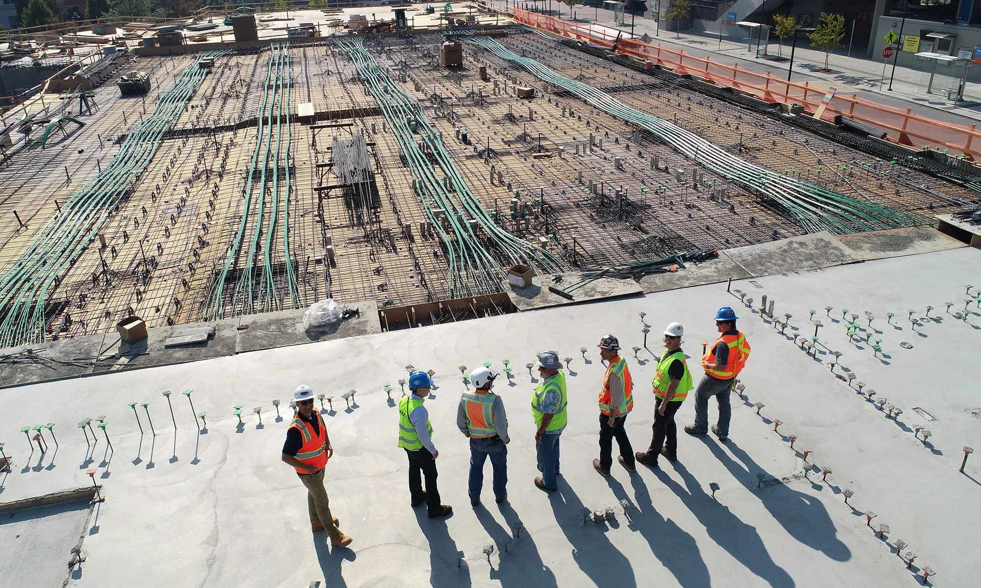 Seven construction workers wearing safety gear standing on a concrete platform near a large construction site.