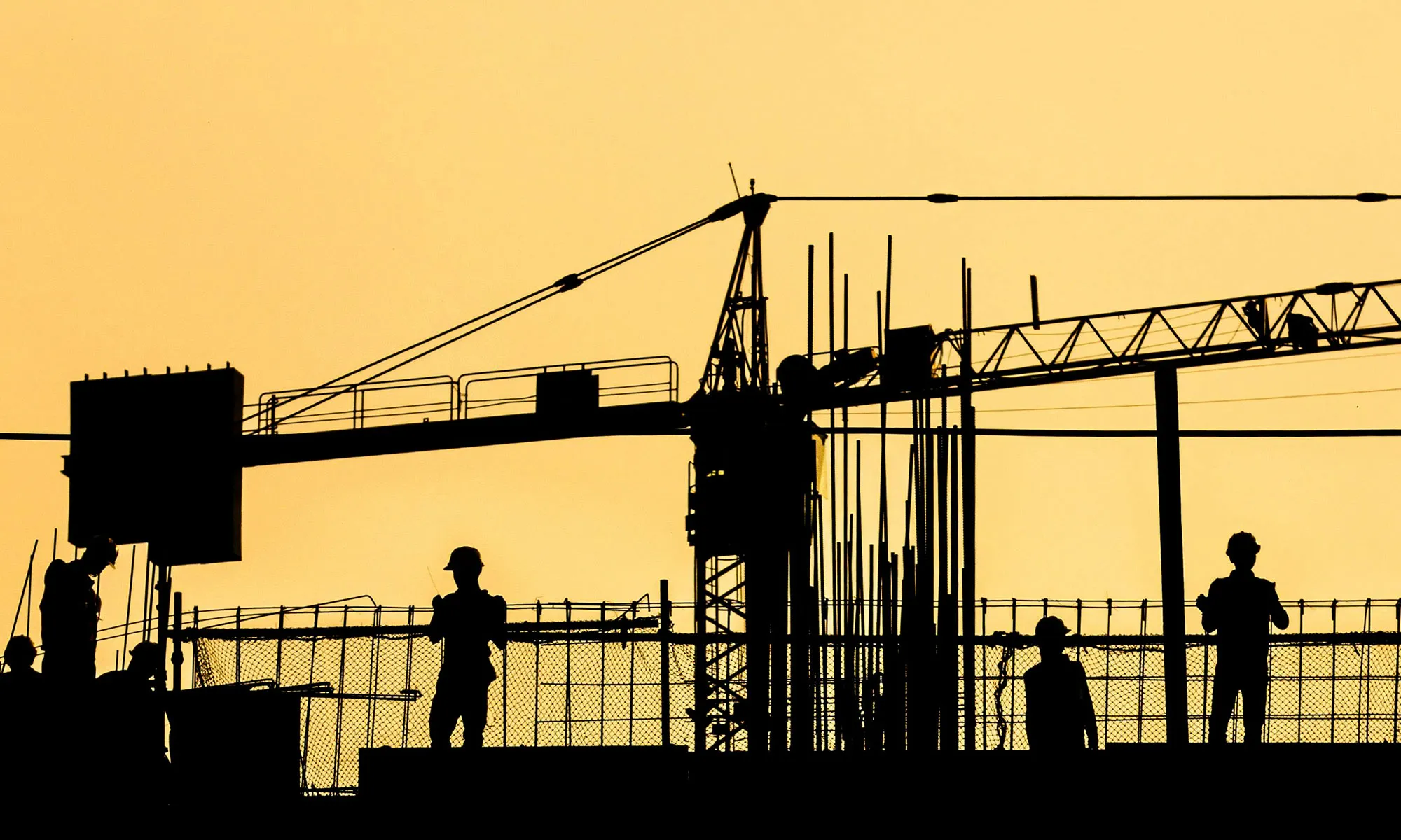 Silhouettes of construction workers and crane structure against an orange sky at sunset.