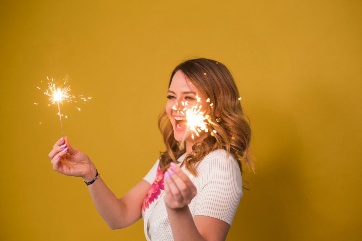 A woman laughing with lit sparklers in both hands against a warm gold background
