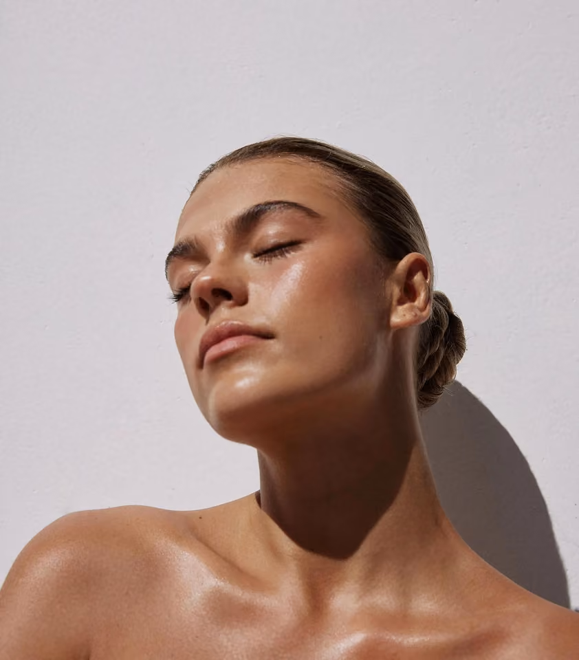 Woman with smooth, glowing skin and eyes closed, basking in sunlight against a plain background.