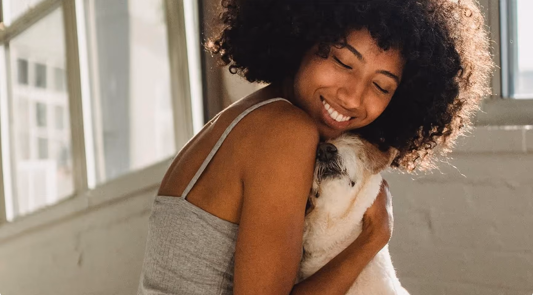 Woman with curly hair smiling and hugging a small dog indoors by a window.