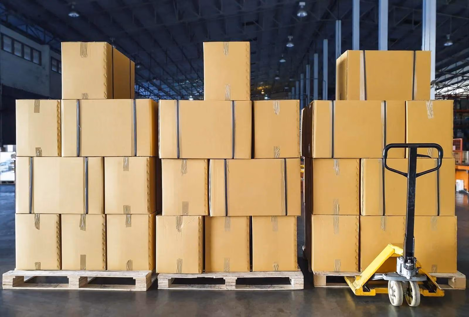 Stacked cardboard boxes on wooden pallets with yellow pallet jack in warehouse interior.