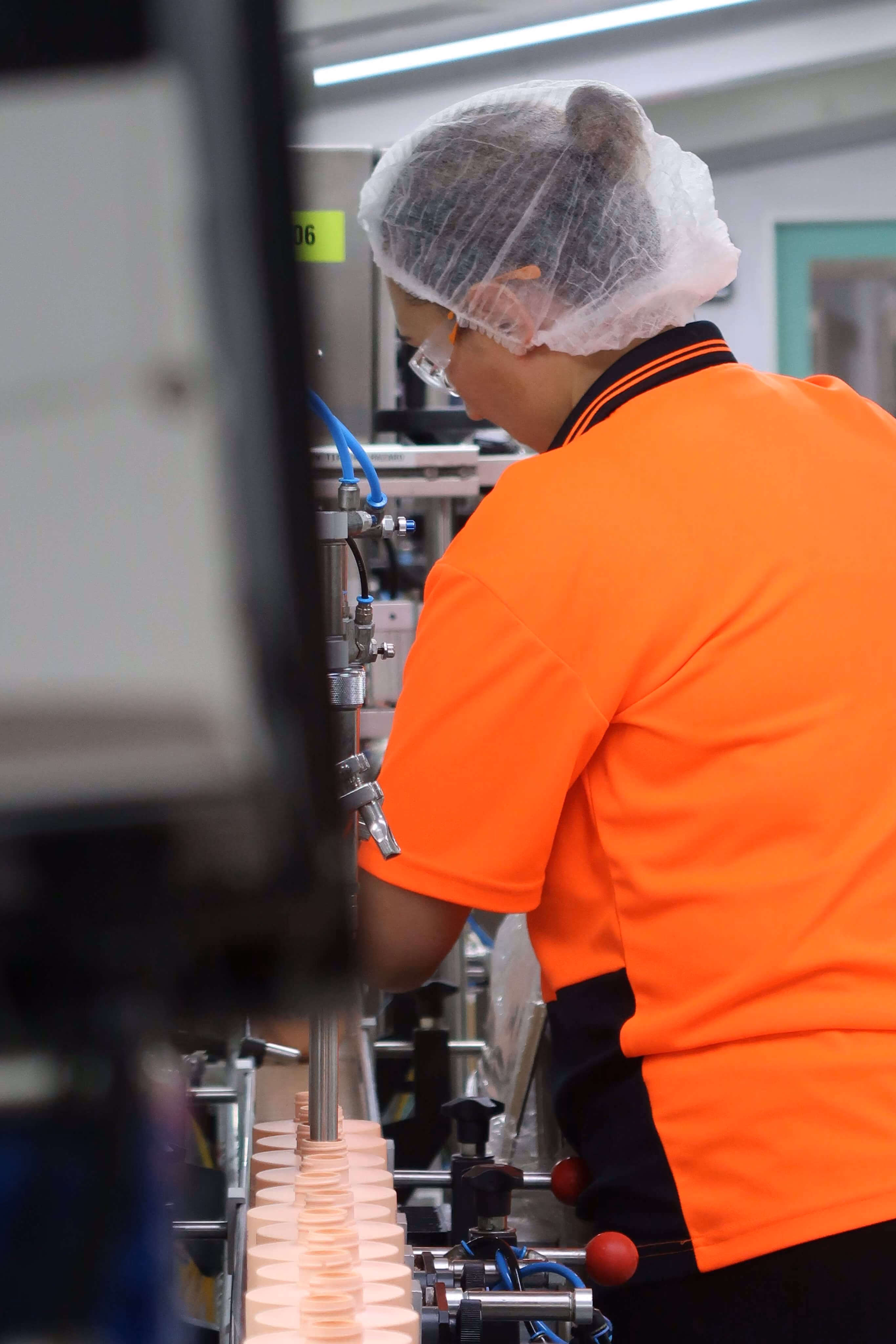 Worker in orange shirt and hairnet operating machinery filling cream containers on a production line.
