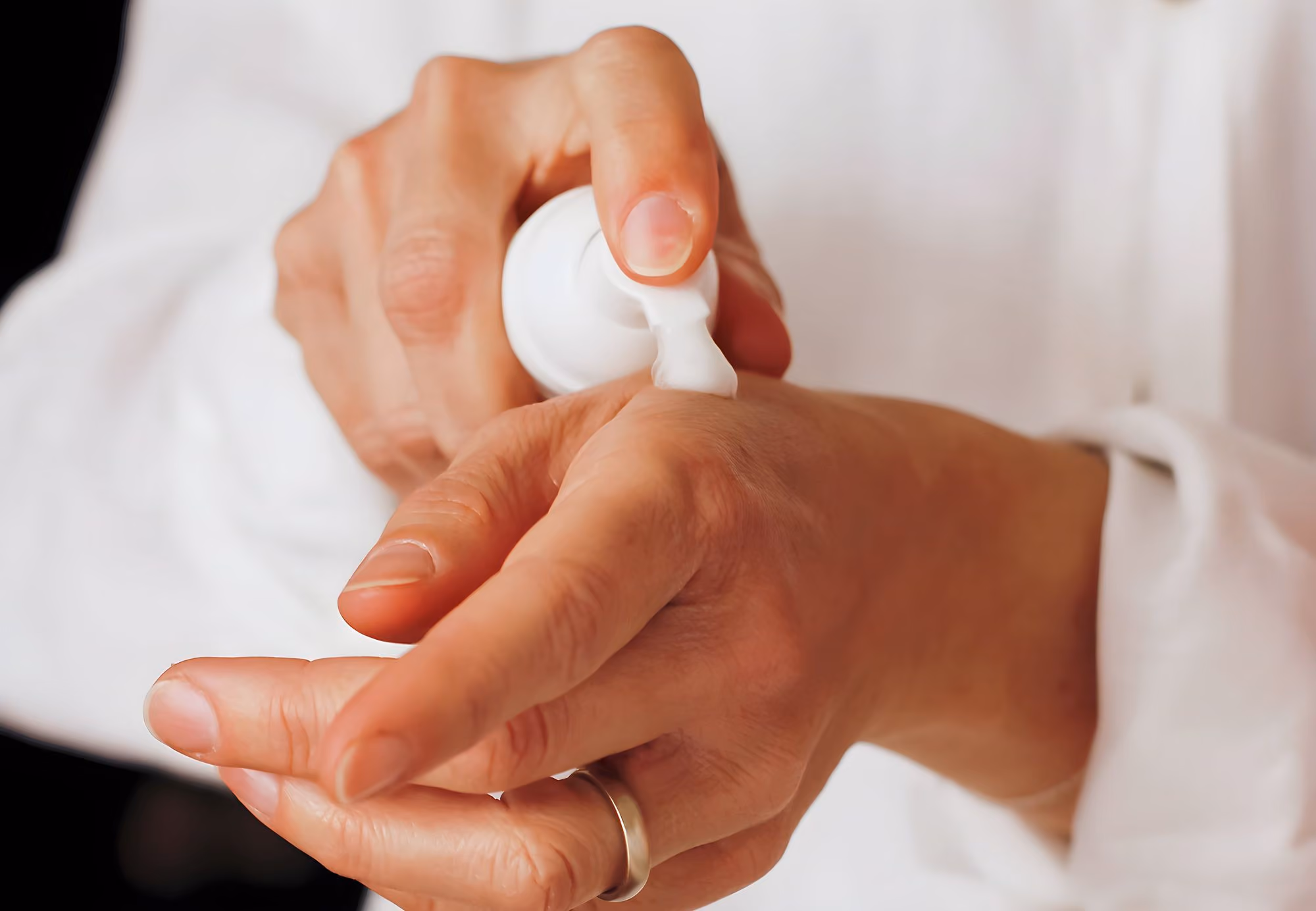 Person wearing a ring applying white cream to the back of their hand from a pump bottle.