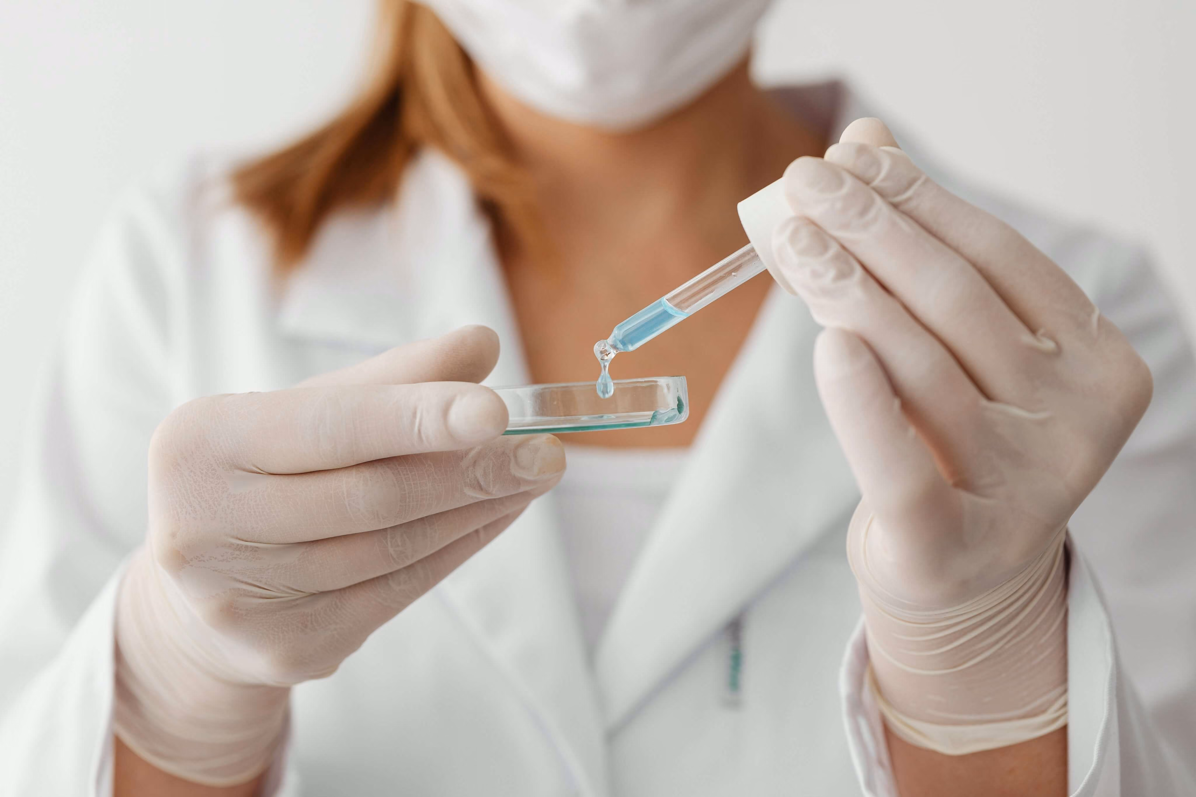 Scientist wearing gloves dropping blue liquid from a pipette into a petri dish.