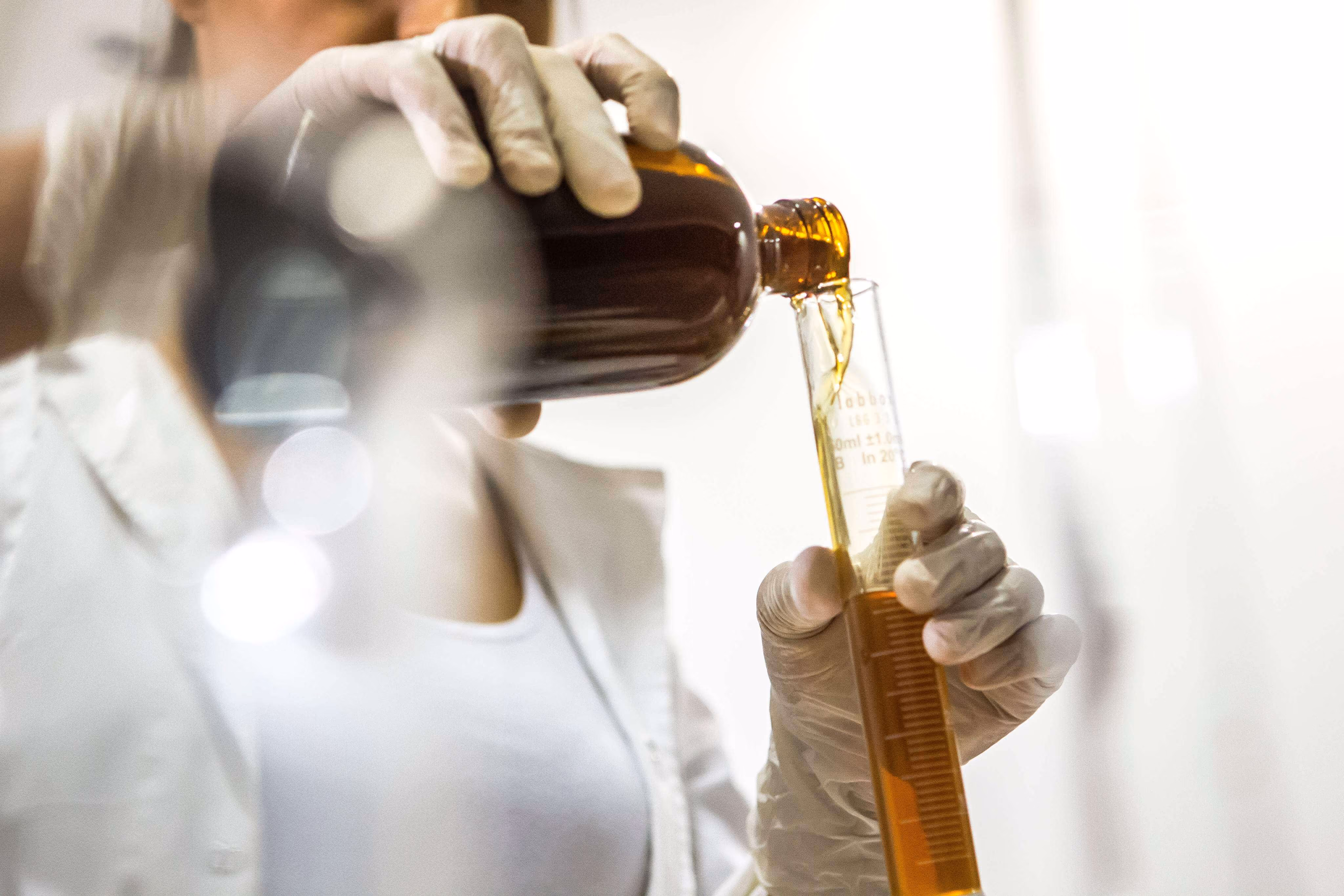 Person wearing gloves pouring amber liquid from a brown bottle into a graduated cylinder in a lab.