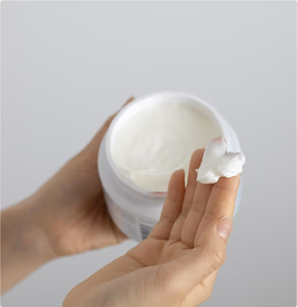 Hand holding white jar and scooping out thick white cream with fingers.