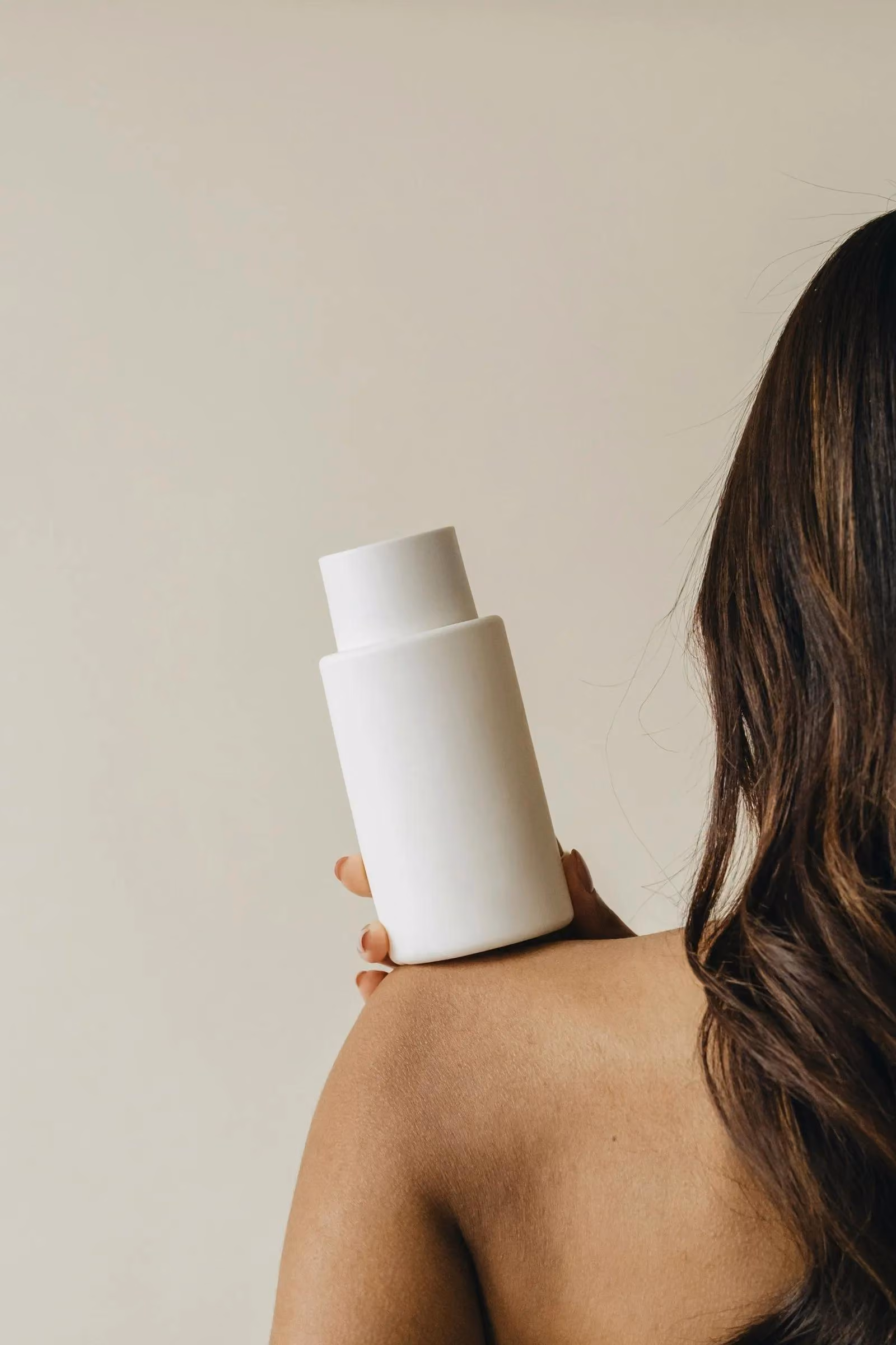 Person with dark wavy hair holding a plain white cylindrical bottle on bare shoulder.