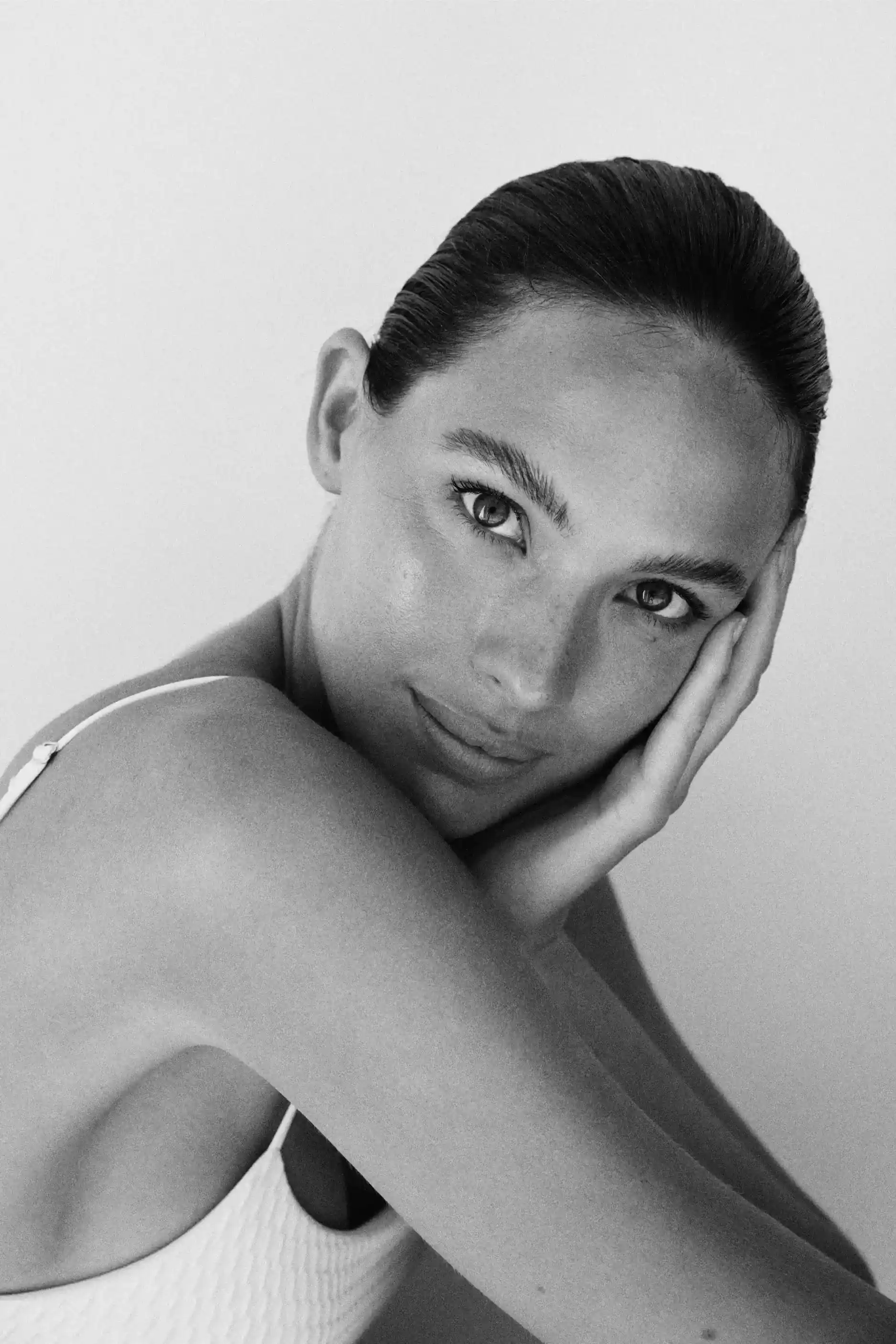 Black and white close-up portrait of a woman with dark hair pulled back, resting her face on her hand, wearing a textured white sleeveless top.