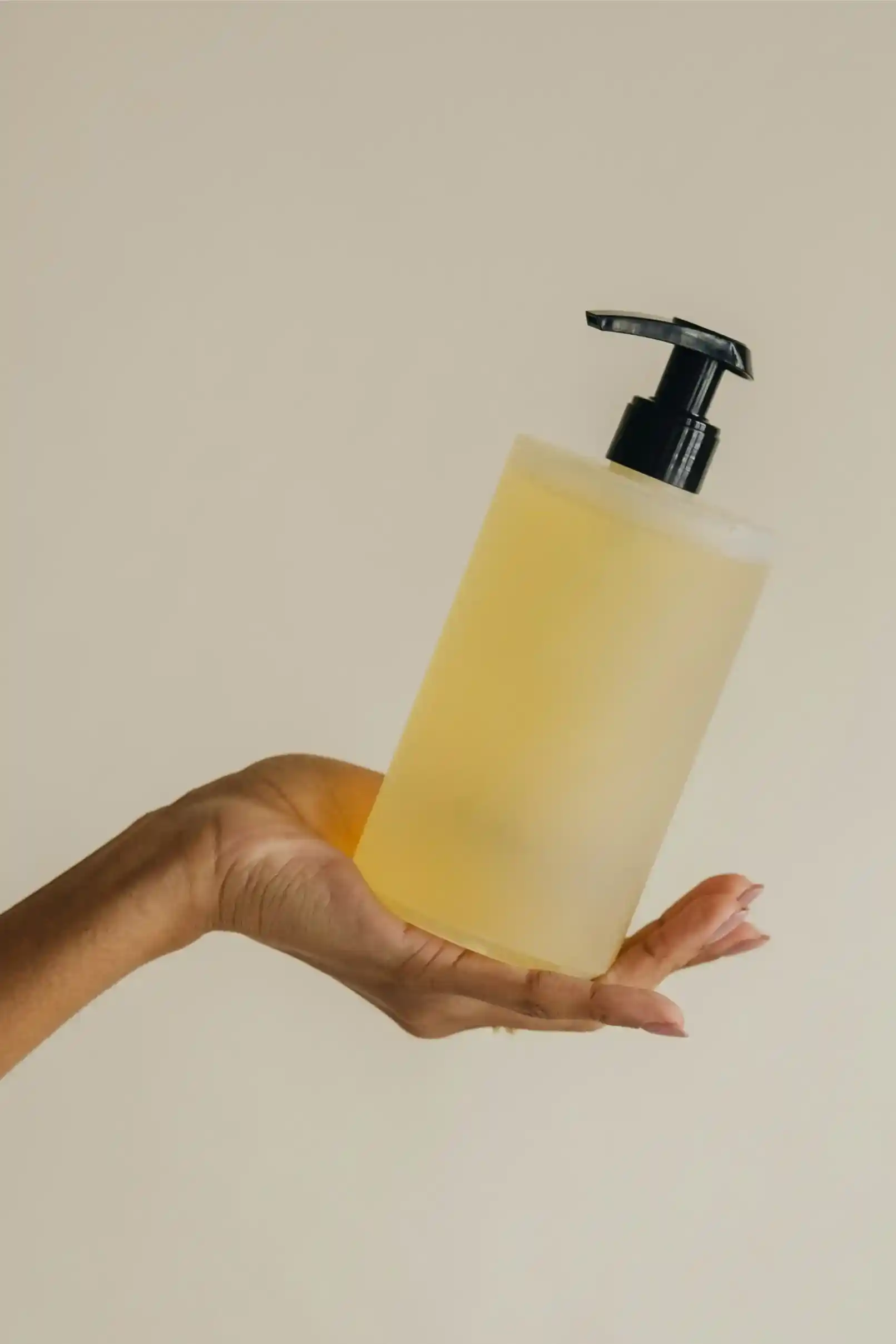A hand holding a translucent yellow liquid soap dispenser with a black pump against a plain beige background.
