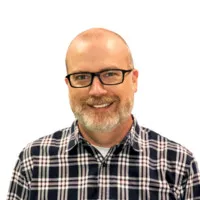 A bald man with light skin, glasses, and a short beard, smiling and wearing a black and white checkered shirt over a white undershirt, against a white background.