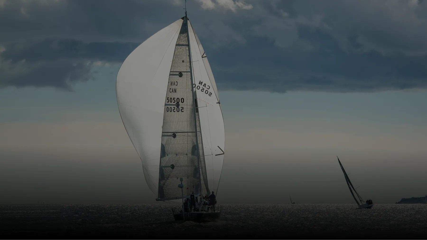 Sailboat with large white sails on calm water under a cloudy sky at dusk or dawn, with a smaller sailboat in the distance.