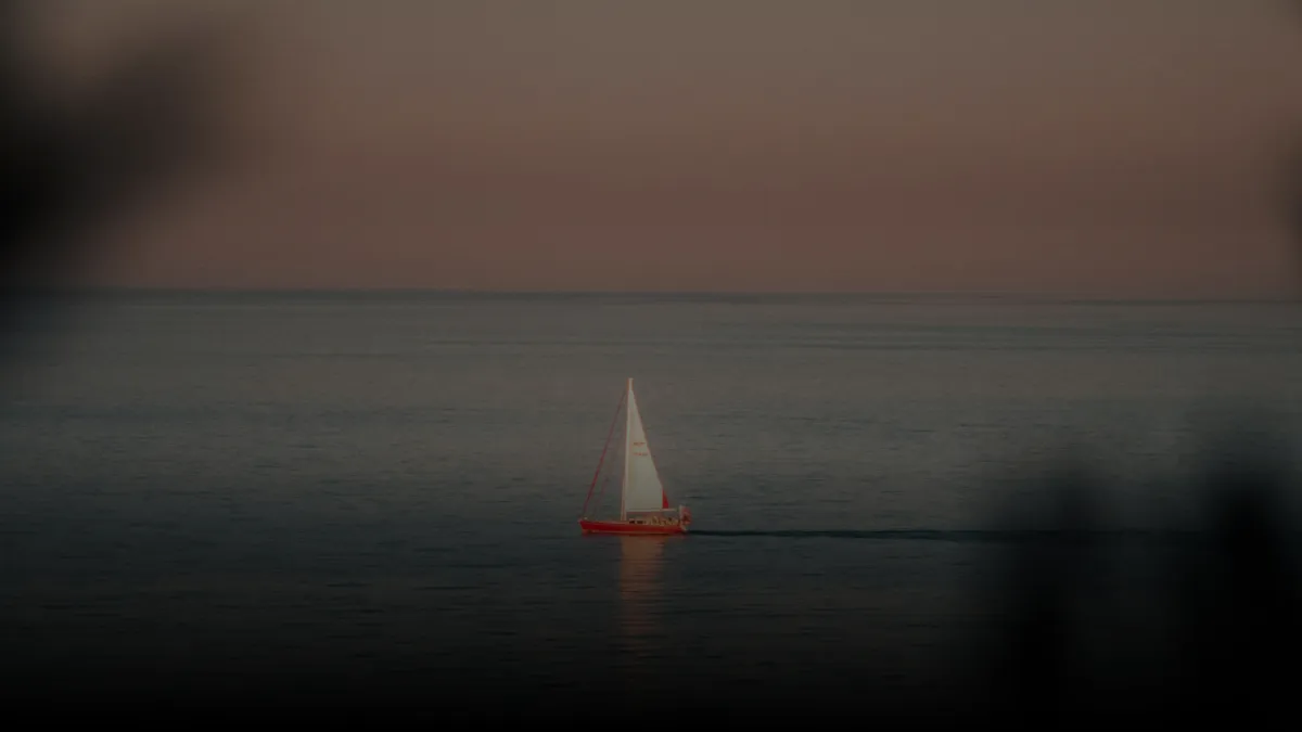 Small red sailboat with white sails gliding on calm water during dusk with a muted pink and gray sky.