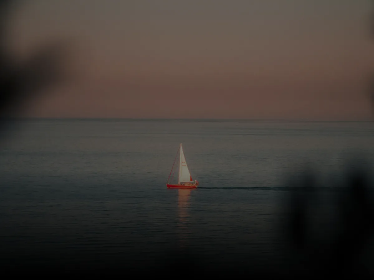 Red sailboat with white sails sailing on calm water at sunset with a pinkish-orange sky.