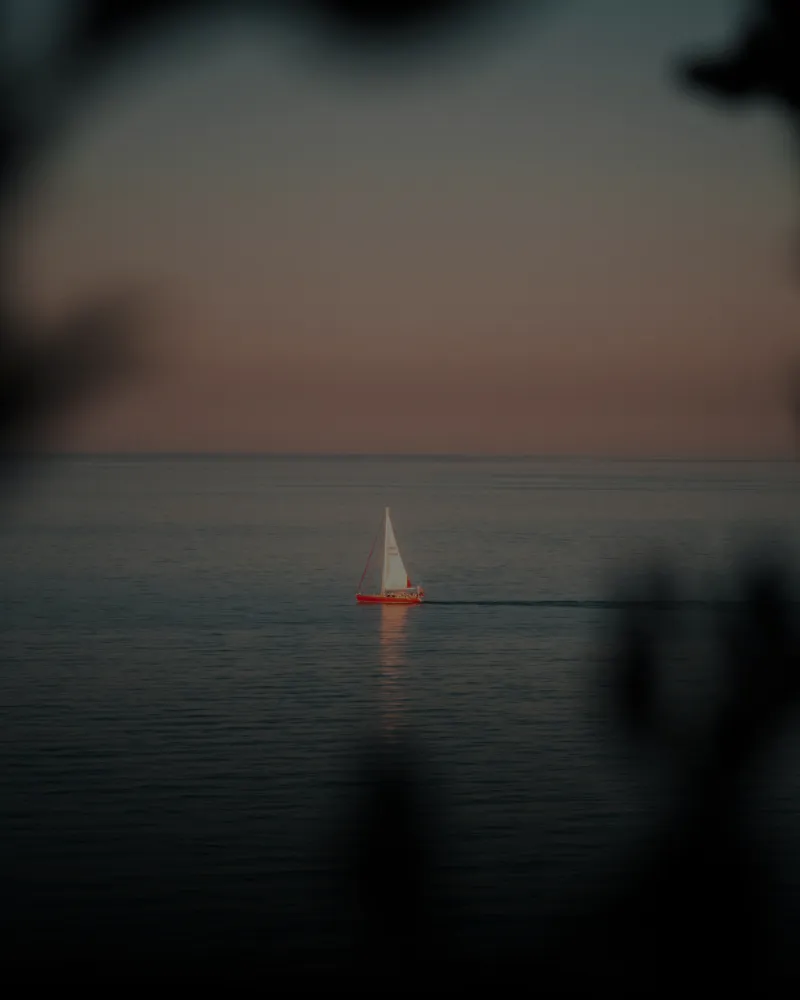 Small sailboat with white sails and red hull sailing on calm water during sunset or sunrise, framed by dark blurred foreground silhouettes.